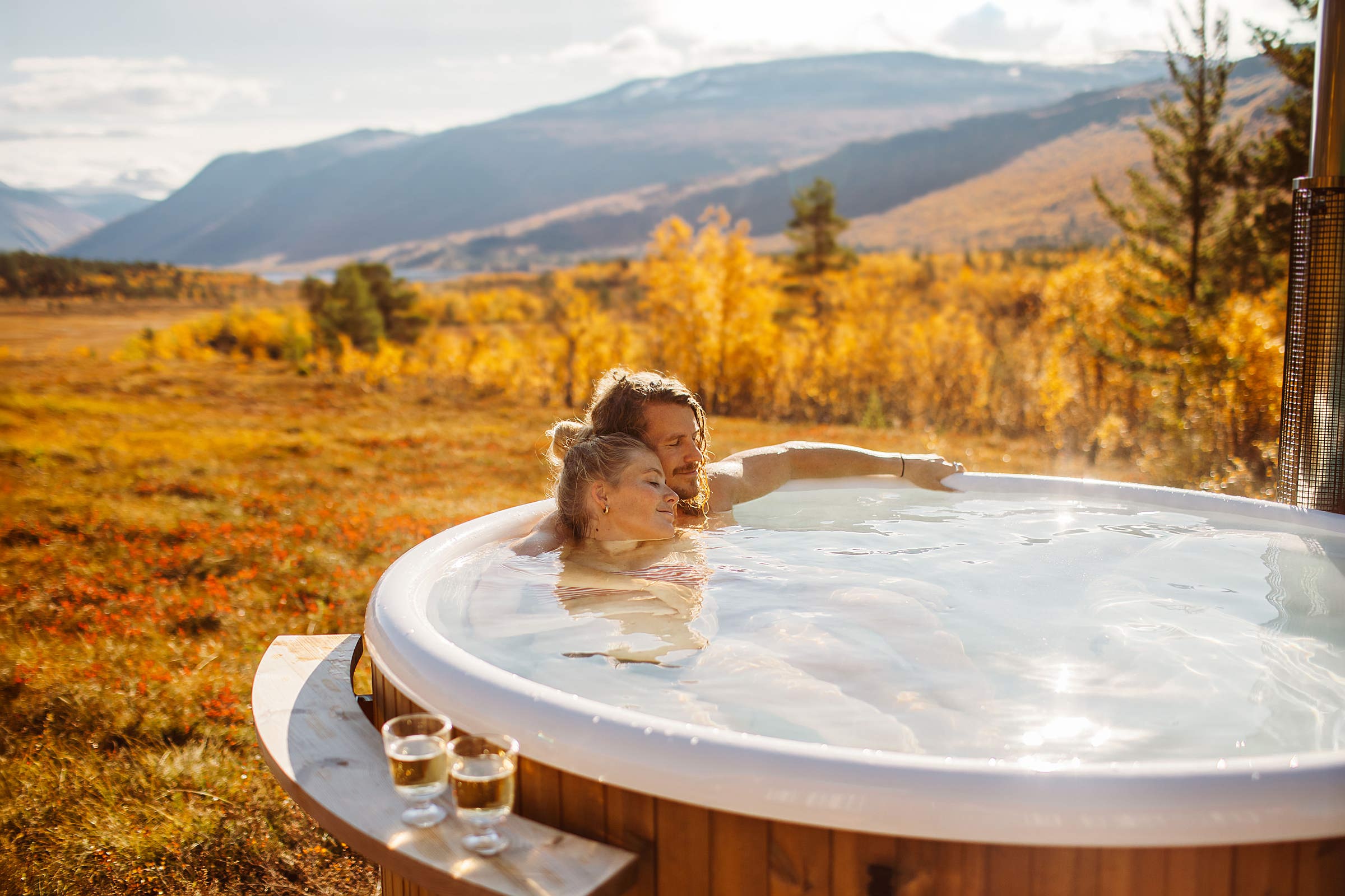 A woman enjoys a wooden hot tub in a field with autumn trees, mountains in the background.