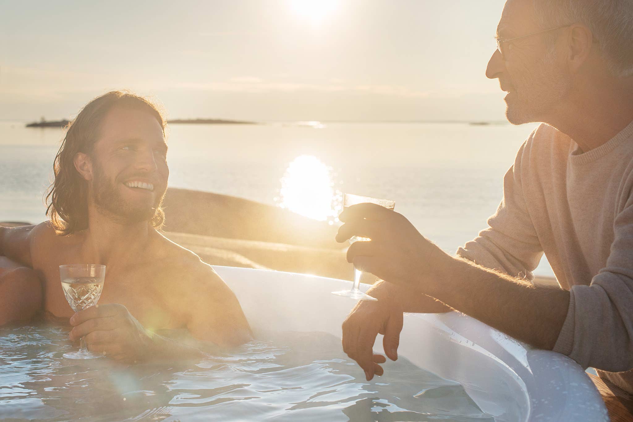 Two men enjoying a Skargards Regal hot tub at sunset with champagne, ocean view, and islands in the background.
