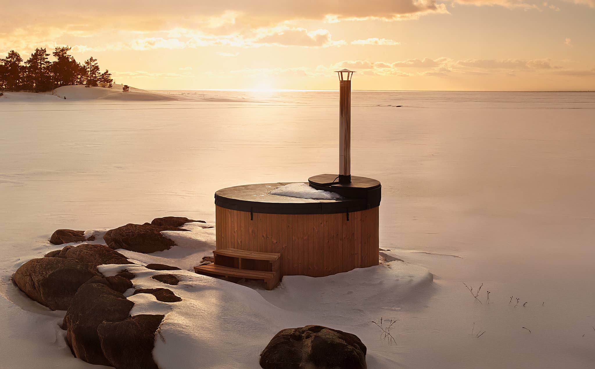 A wooden hot tub with a black insulated lid on a snowy island at sunset.