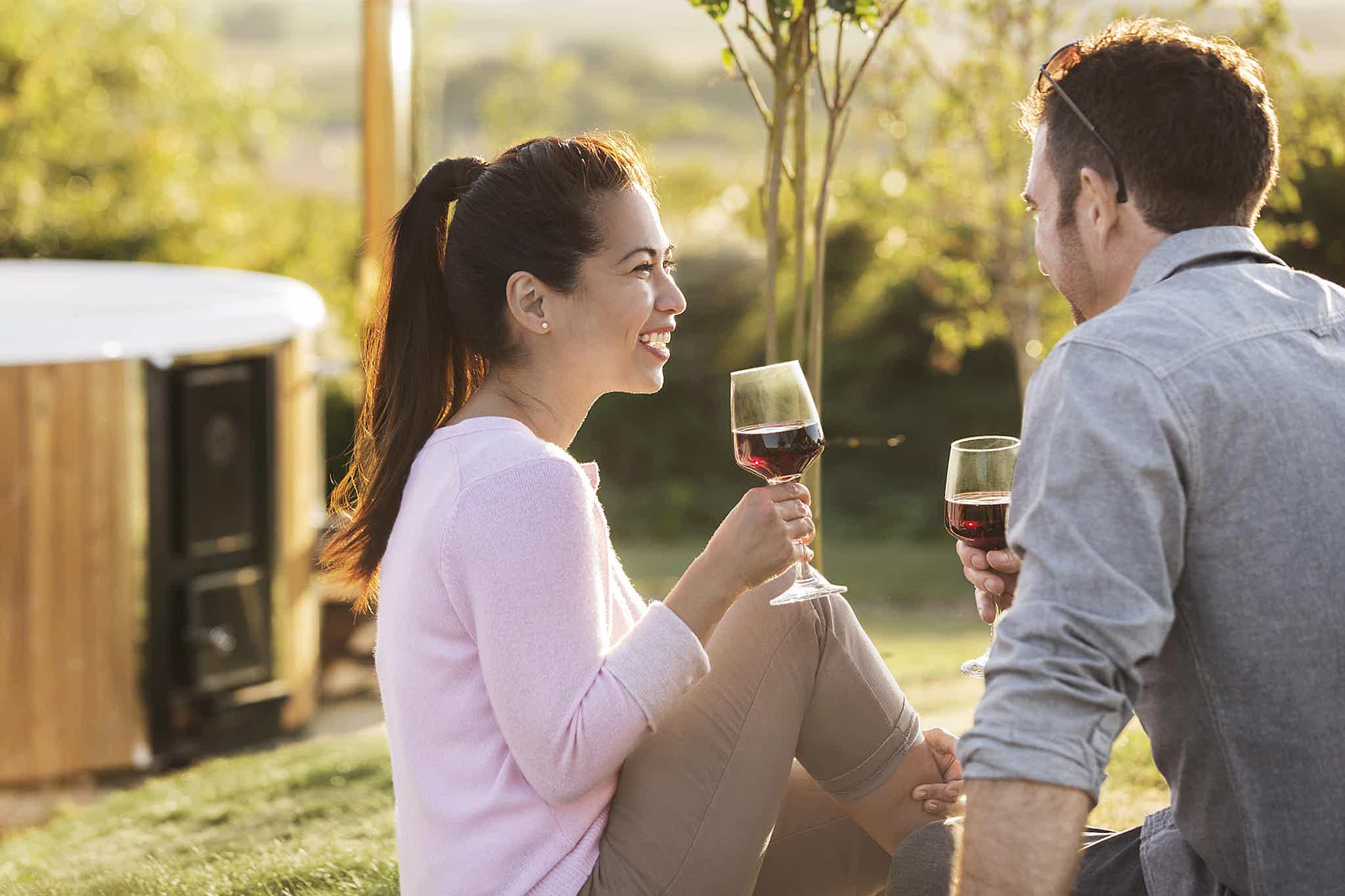 A couple enjoys wine outdoors near a Skargards wood-fired hot tub, surrounded by nature's tranquility.