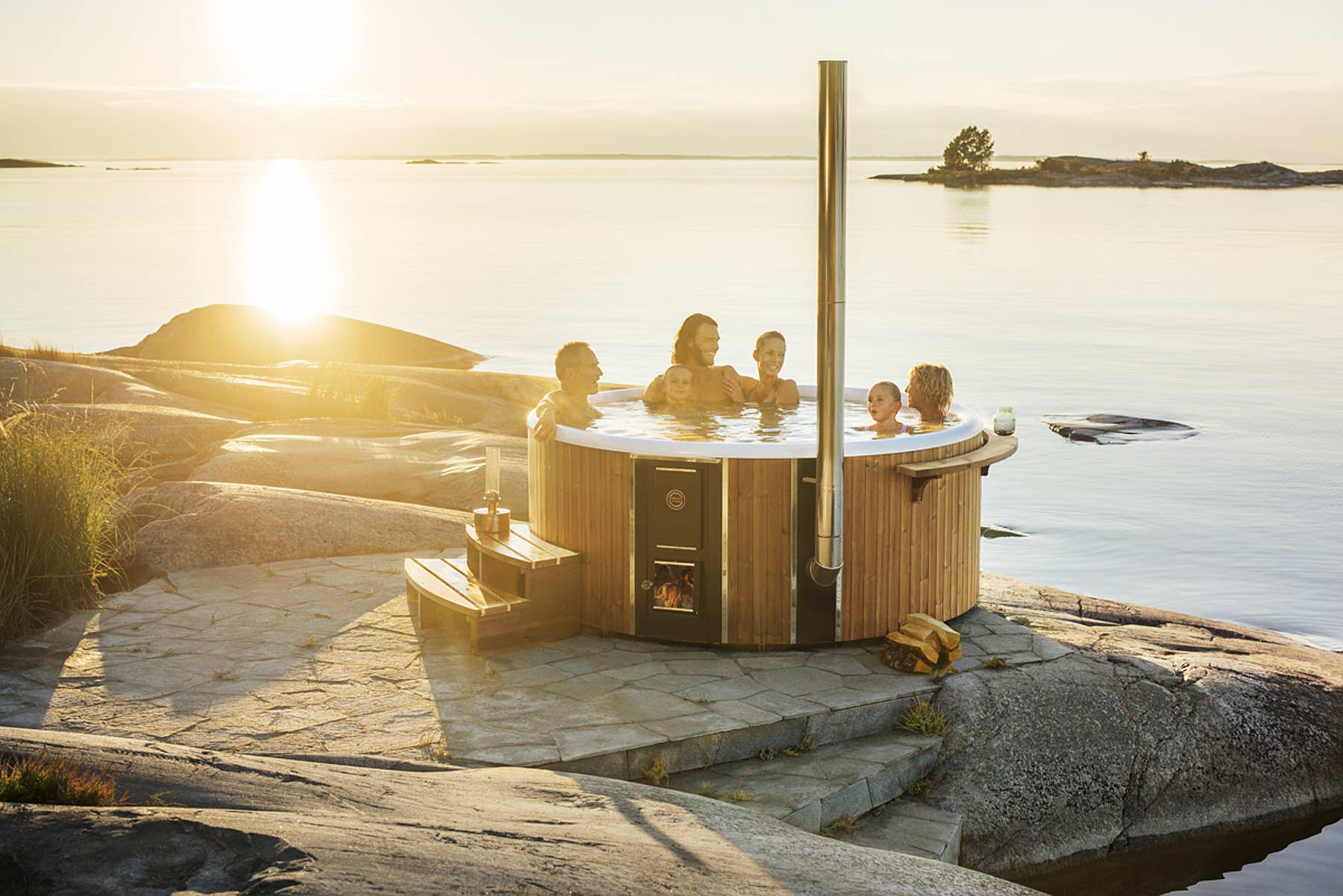 A luxurious wood-fired Skargards Regal hot tub on rocky shore with ocean view. A family enjoys some time together in the warm water of the tub.