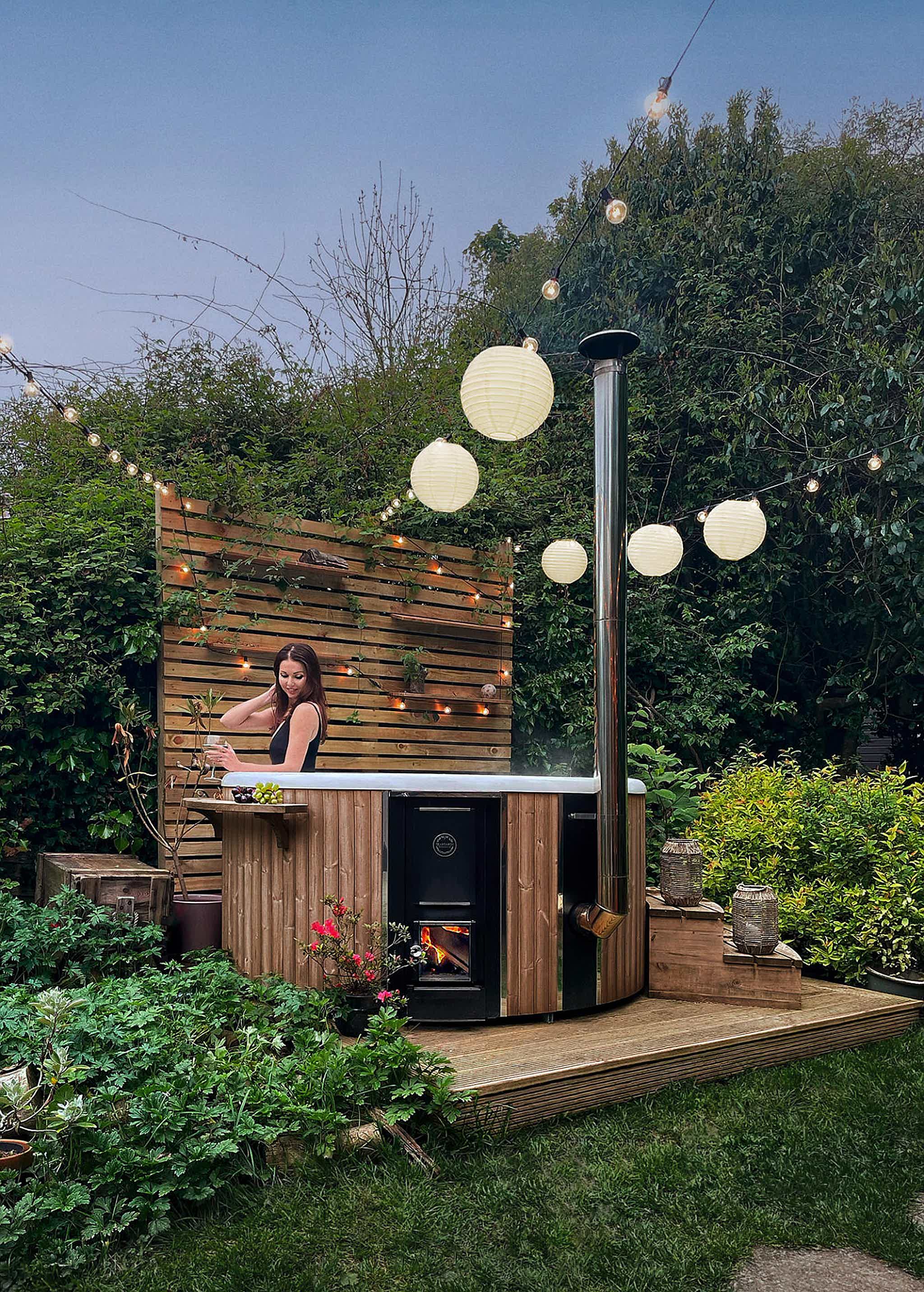 A woman in a black bathing suit in a Skargards Regal 190 hot tub, surrounded by a lush garden and string lights.