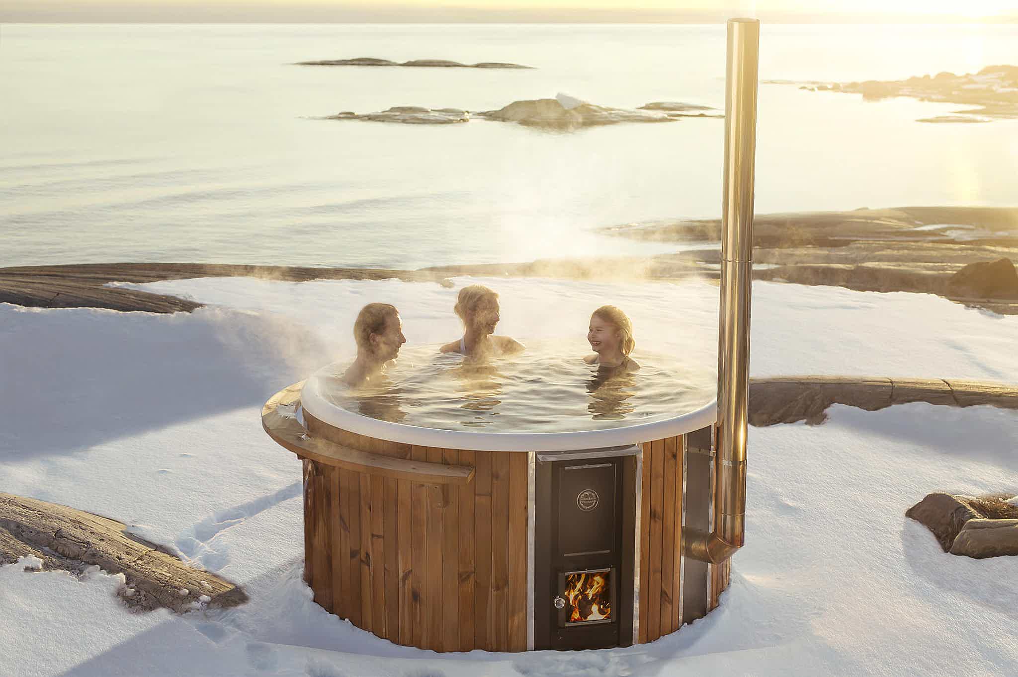 A wood-fired Skargards Regal hot tub in snowy landscape by ocean, a family enjoys warmth and ambiance.