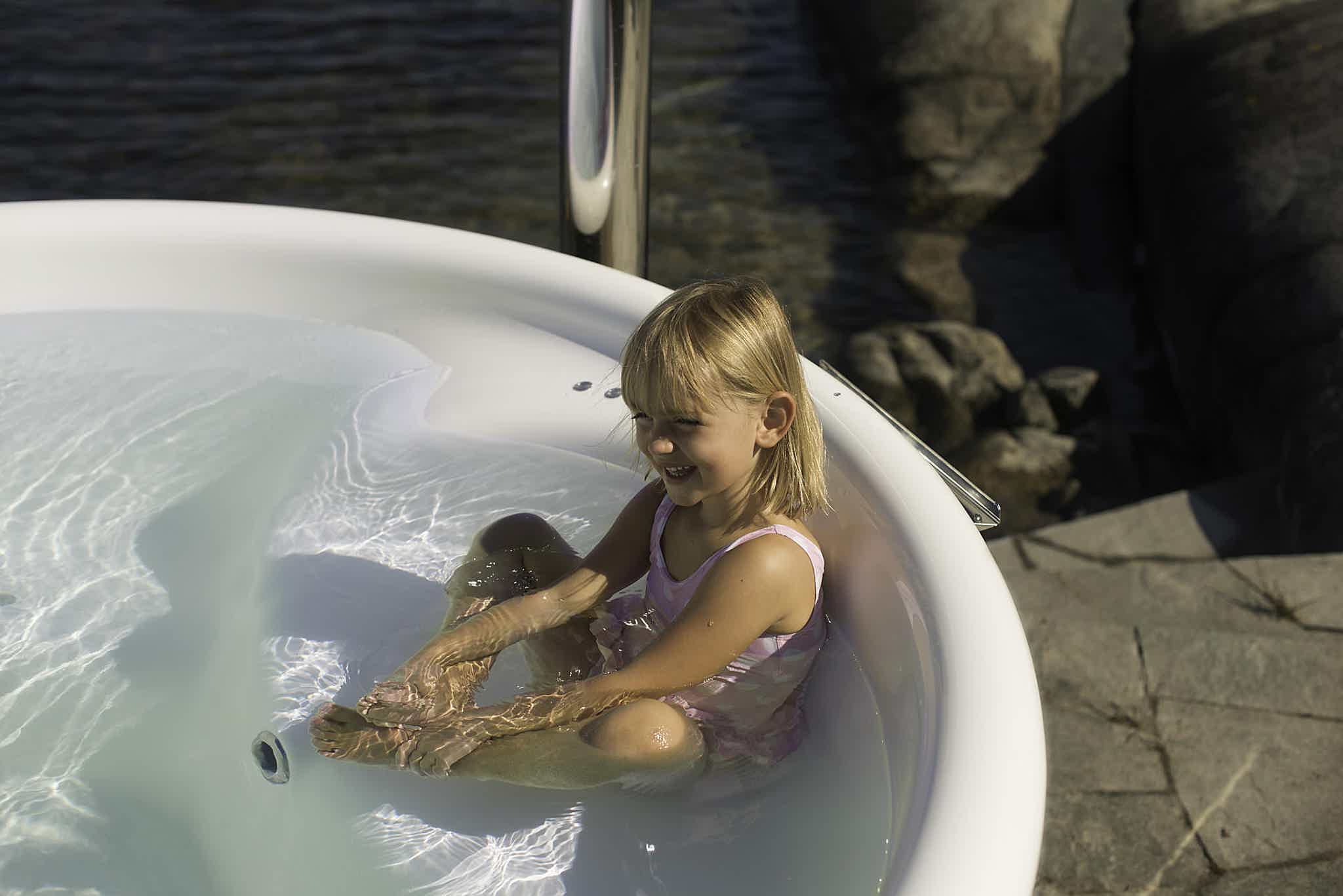 A young girl in a pink swimsuit relaxes in a Skargards Regal hot tub, surrounded by rocks and calm water.
