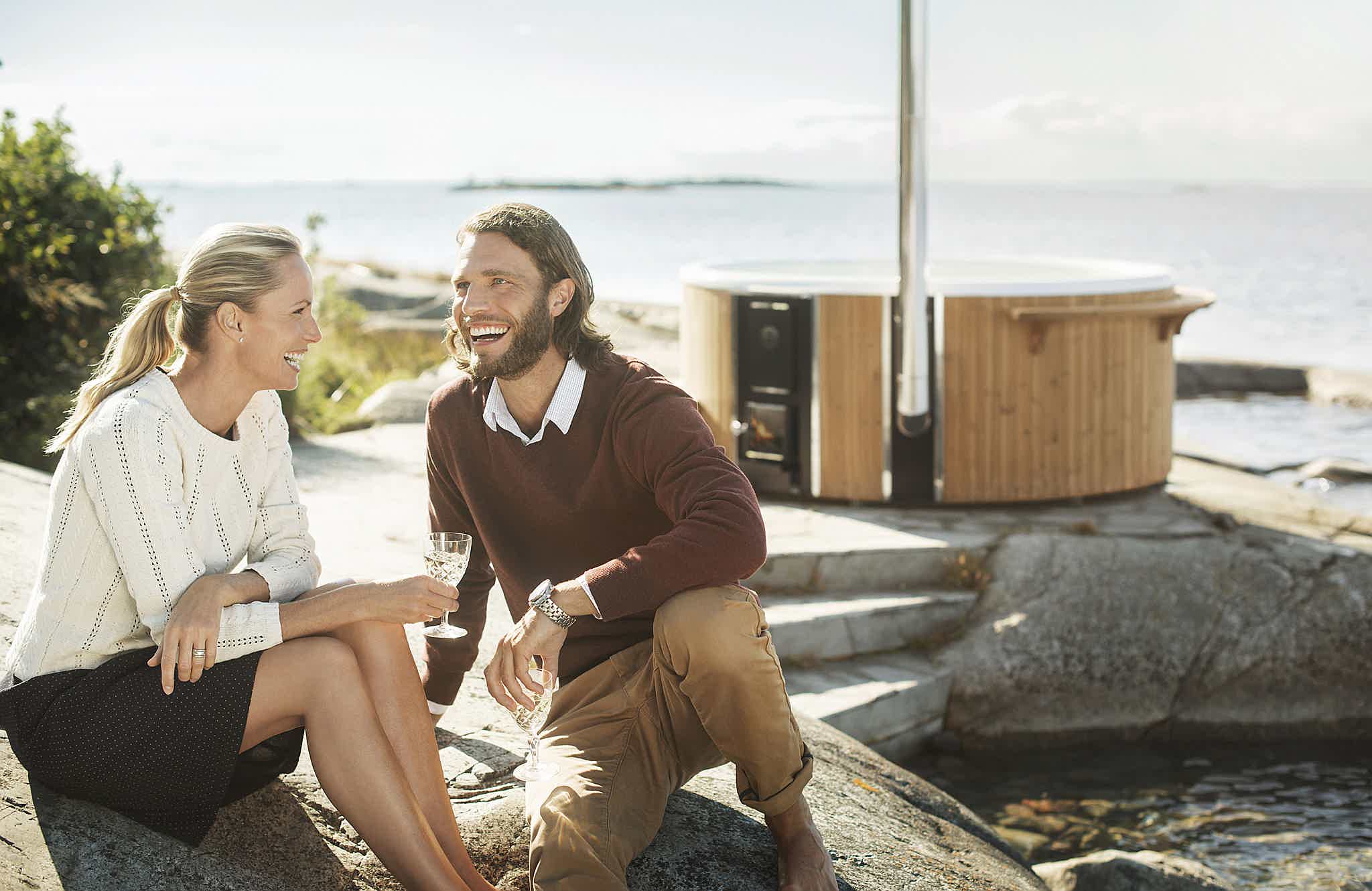 A couple sits by the water with a wood-fired Skargards hot tub in the background, enjoying a clear day.