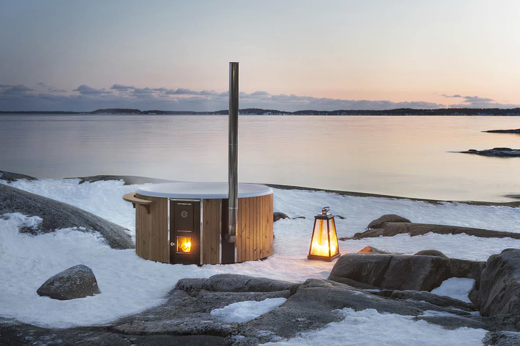 A wood-fired Skargards hot tub on a snowy shore with lanterns, rocks, and water under a dawn or dusk sky.
