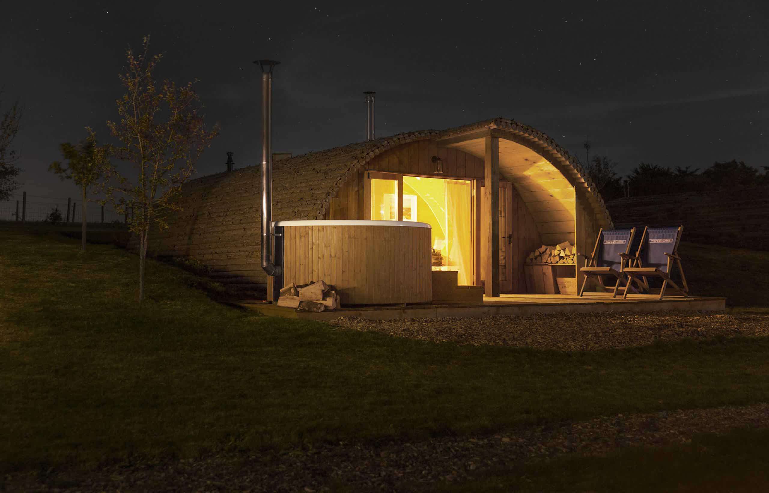 A cabin with a wooden Skargards hot tub and chairs on a hill, glowing warmly at night.