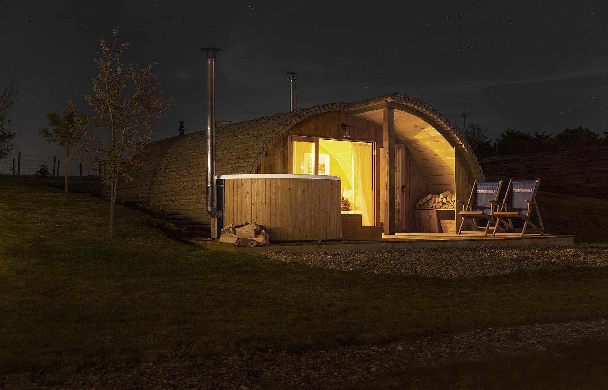 A cabin with a wooden Skargards hot tub and chairs on a hill, glowing warmly at night.