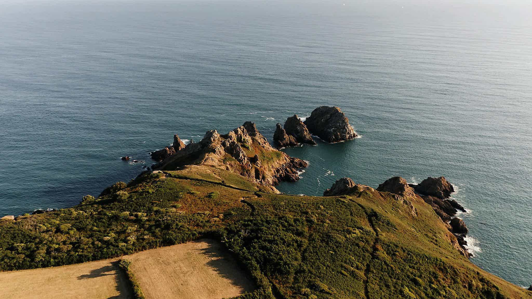 Aerial view of the Jerbourg Point on Guernsey, a rocky coastline with calm blue water and grass-covered land.