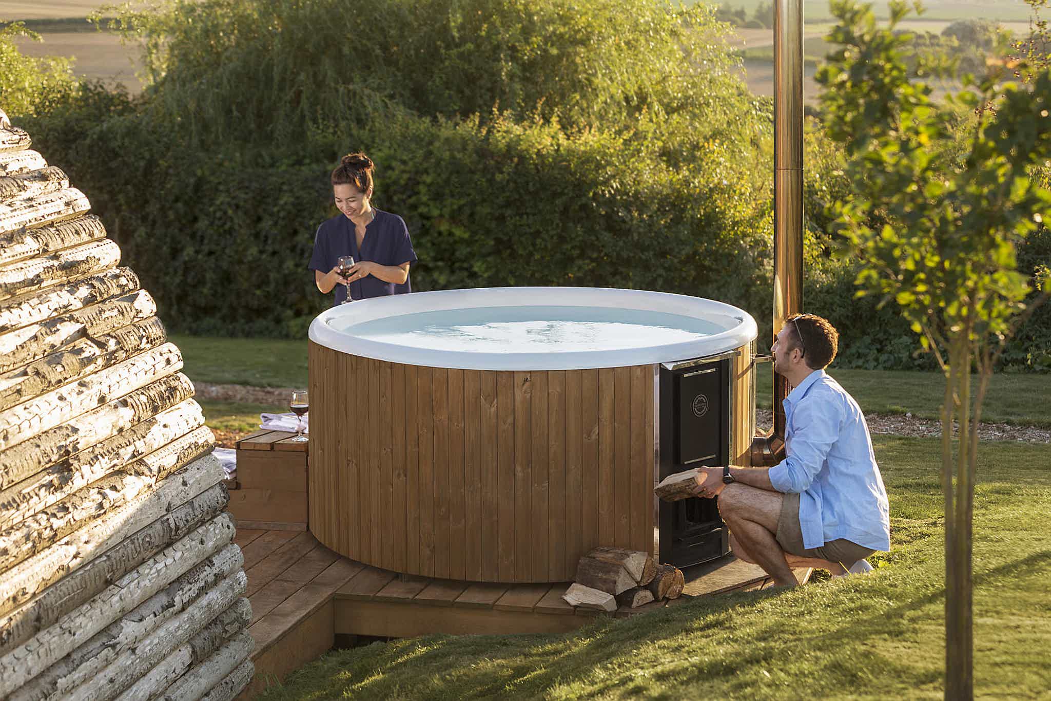 A man kneels by a wooden Skargards Regal hot tub with a log, a woman stands beside him in a lush garden with trees.