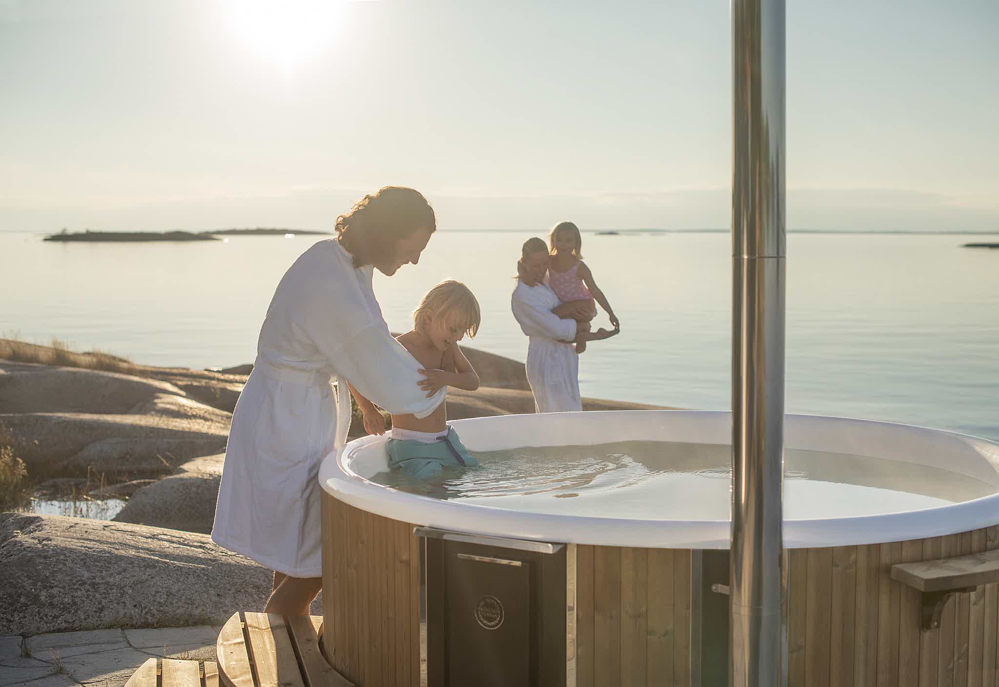 A family in white bathrobes approach a wood-fired Skargards hot tub on a rocky shore to take a bath.