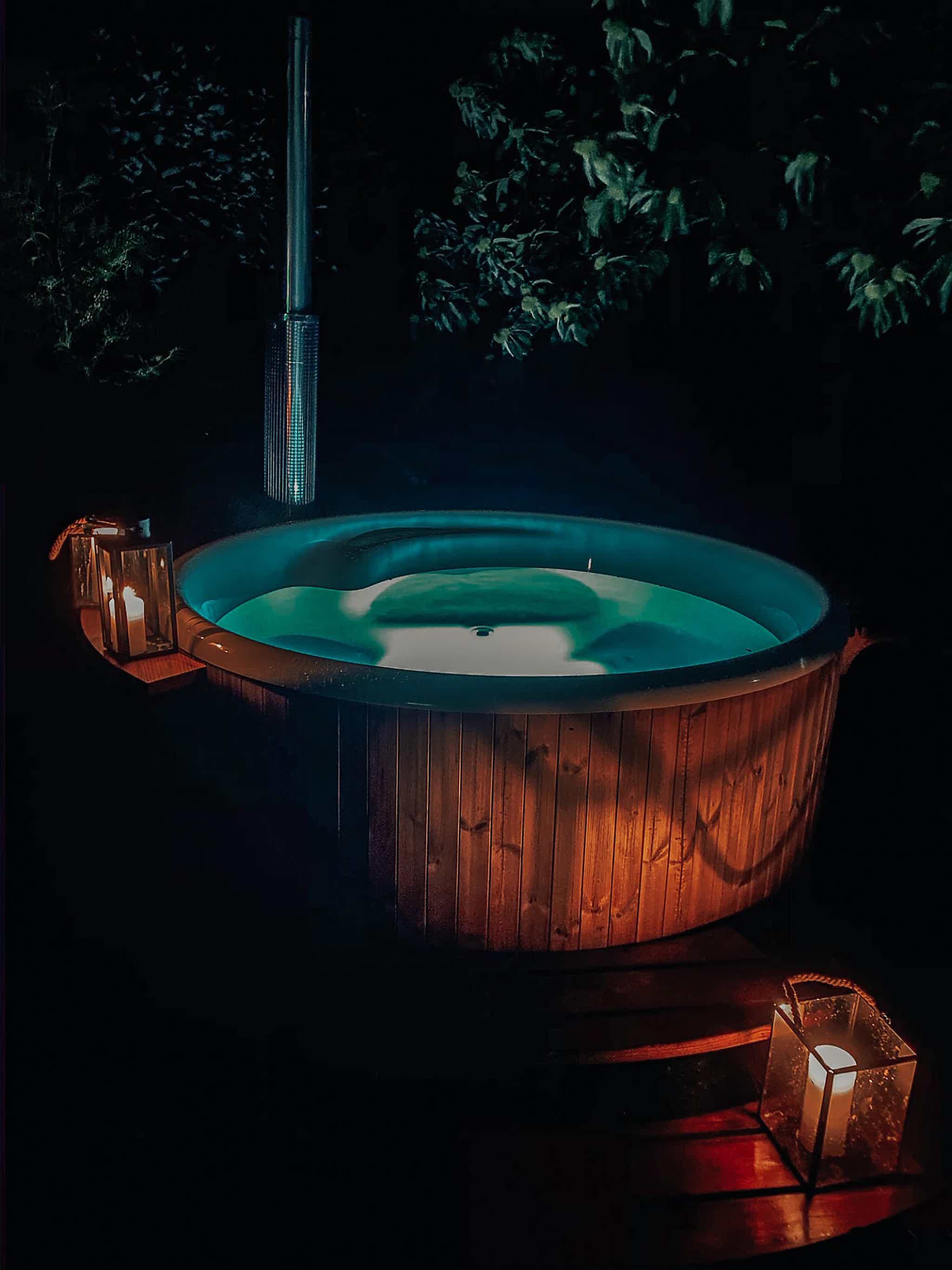 A wooden Skargards Regal hot tub on a platform, lit by a lamp and candle, surrounded by nature for a serene atmosphere.
