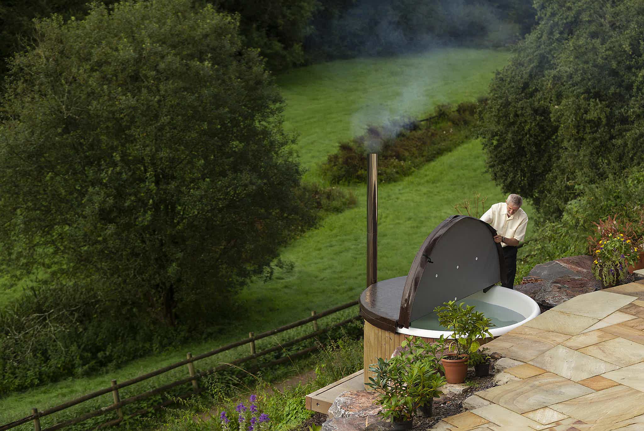 Hugh lifts the insulated lid of his Skargards Regal DualBurn 190 hot tub. Steam rises, with meadows and trees behind.