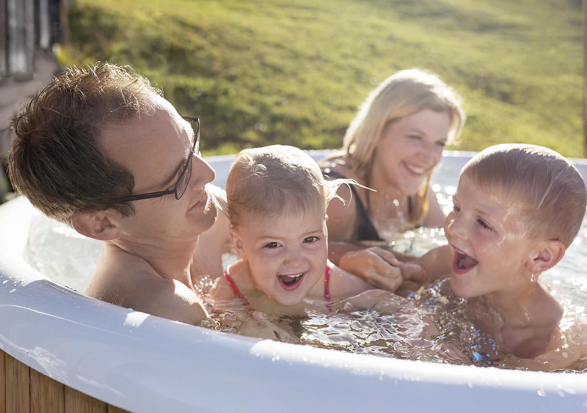 A family enjoys a Skargards hot tub on a green meadow, father with glasses holding laughing kids.