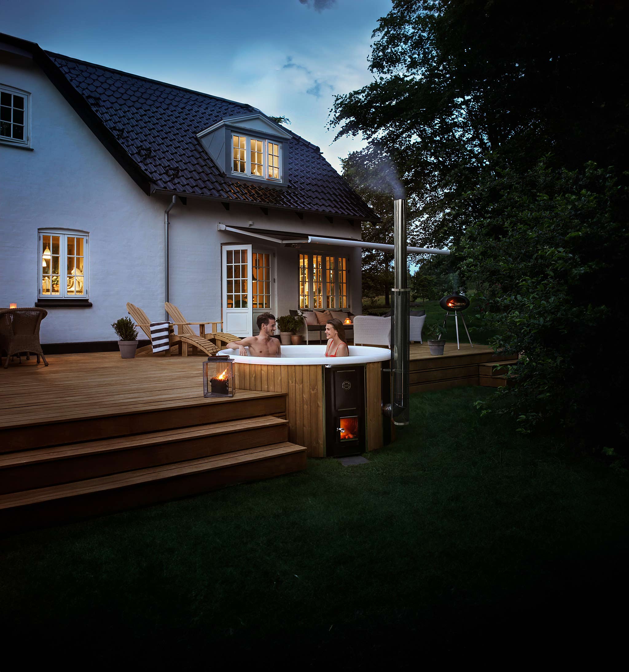 Couple in a hot tub on a wooden deck with candles and fireplace, house with white exterior and black roof.