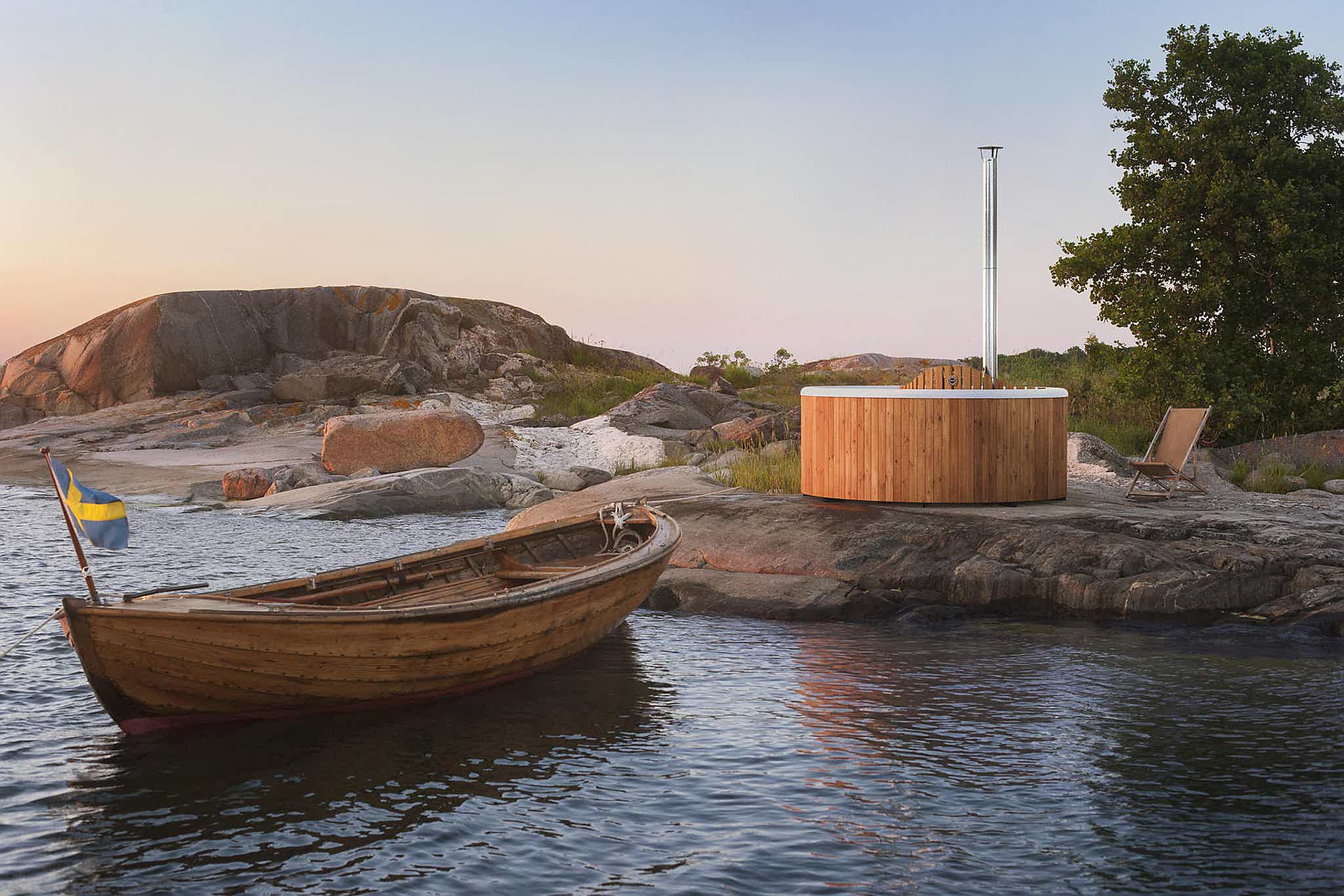 A peaceful lakeside with a wooden boat, hot tub, and flag. Rocks, tree, and orange-pink sky complete the scene.