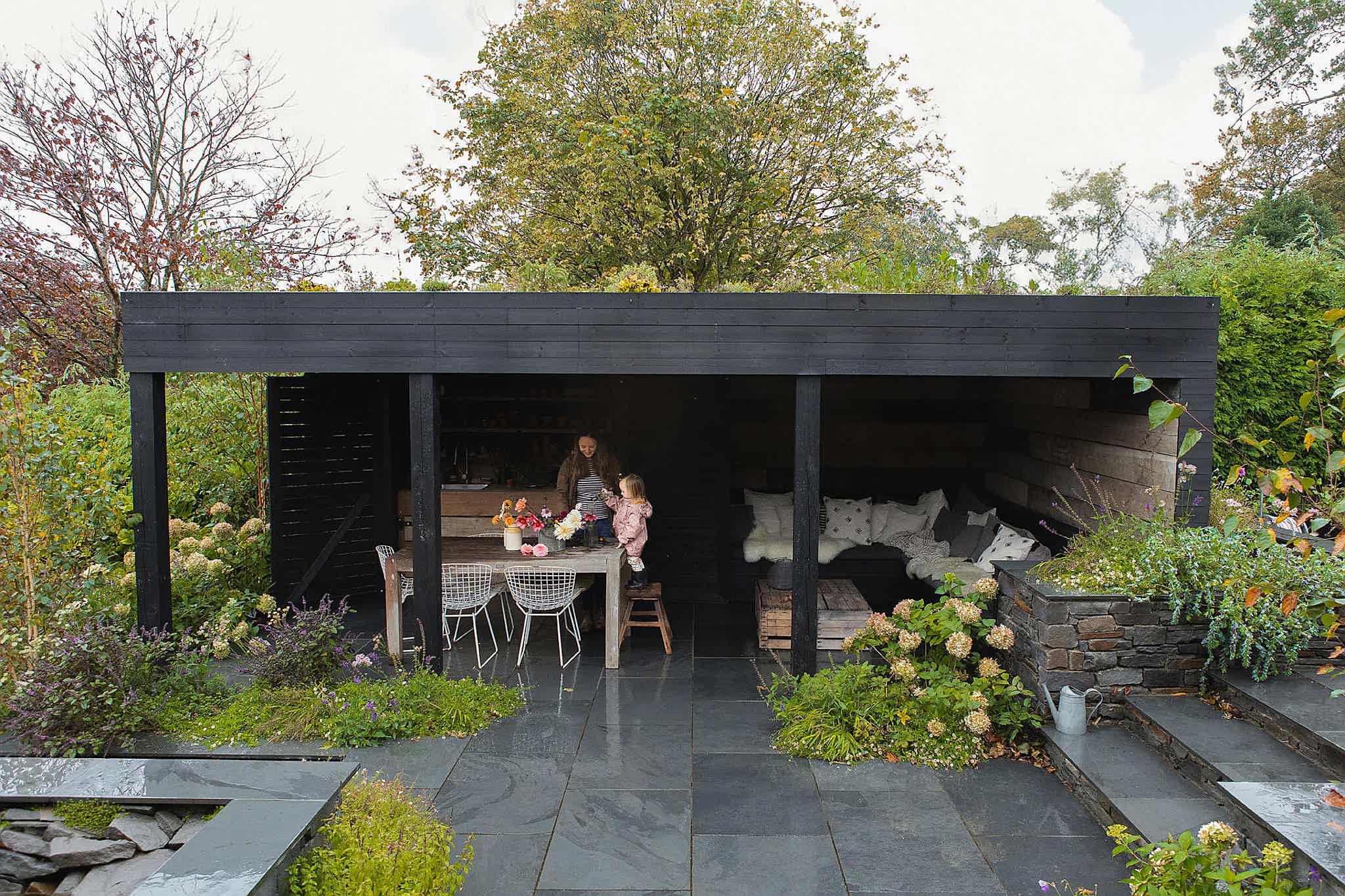 A black wooden seating area with a green roof in a lush garden. Victoria and child arrange flowers on a table.