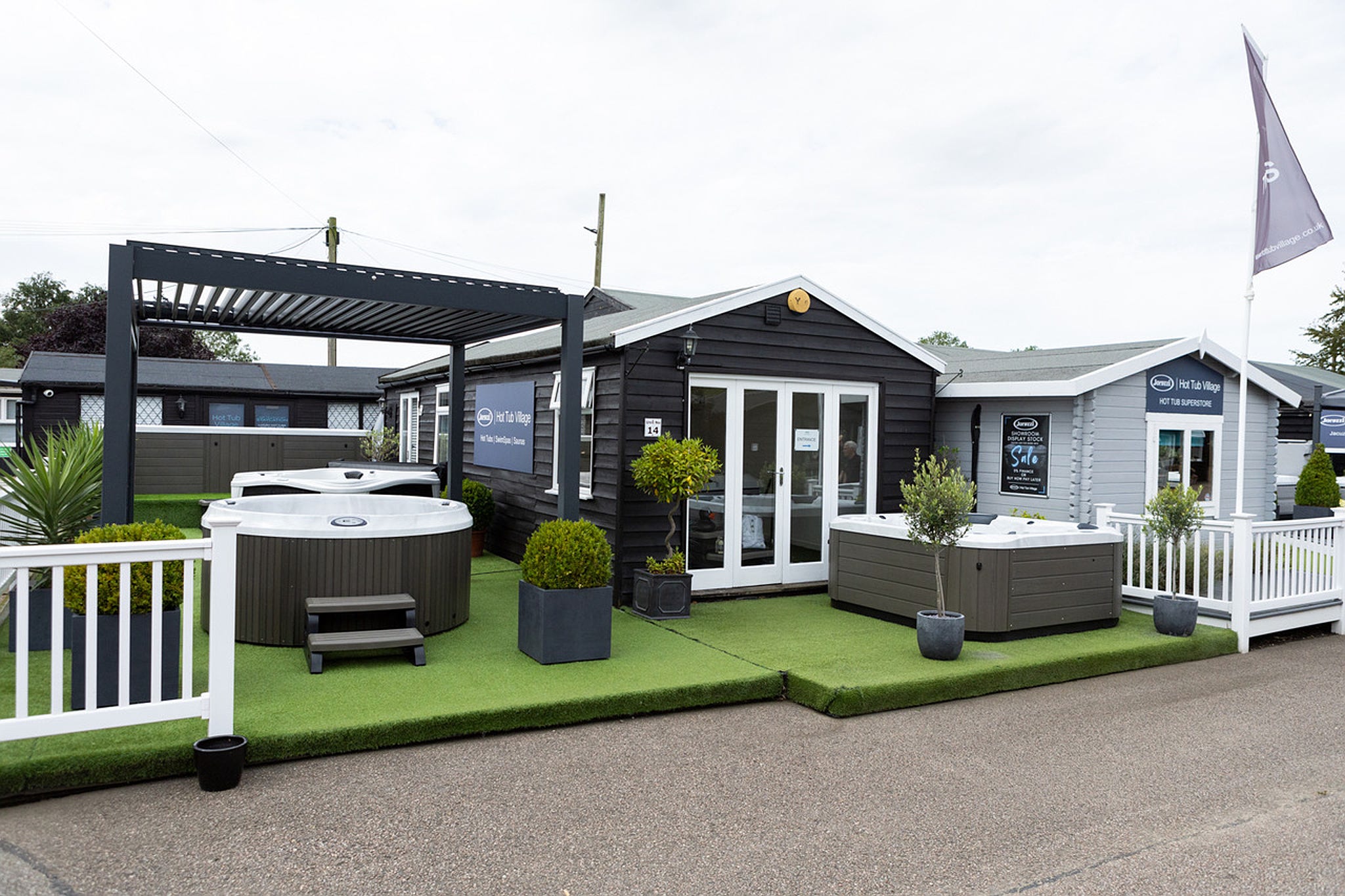 A modern building with a white roof and a hot tub in a green area, surrounded by a white fence and a blue flag.