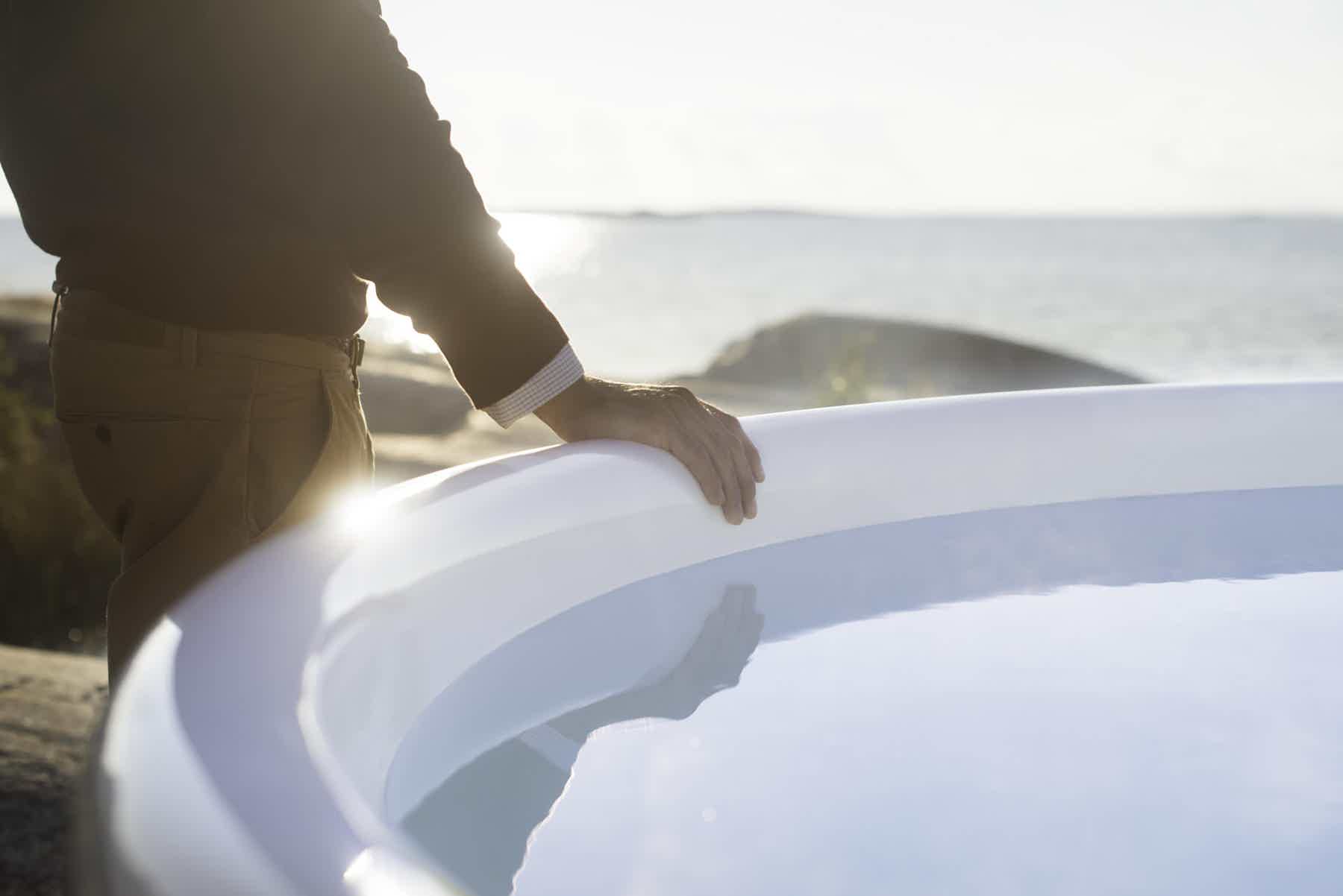 A man rests his hand on the vat edge of a Skargards Regal hot tub under the sun near water.