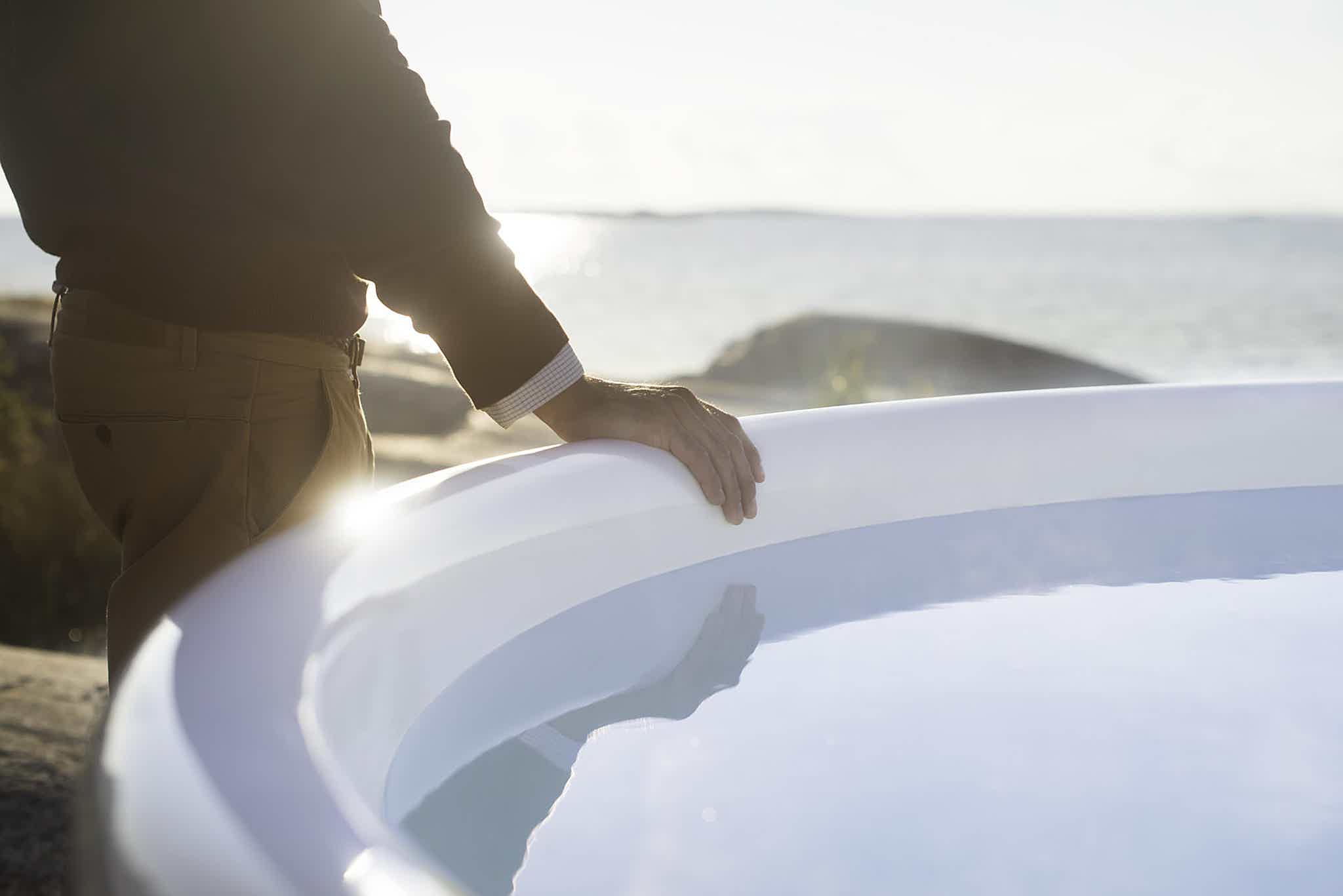 A man rests his hand on the vat edge of a Skargards Regal hot tub under the sun near water.
