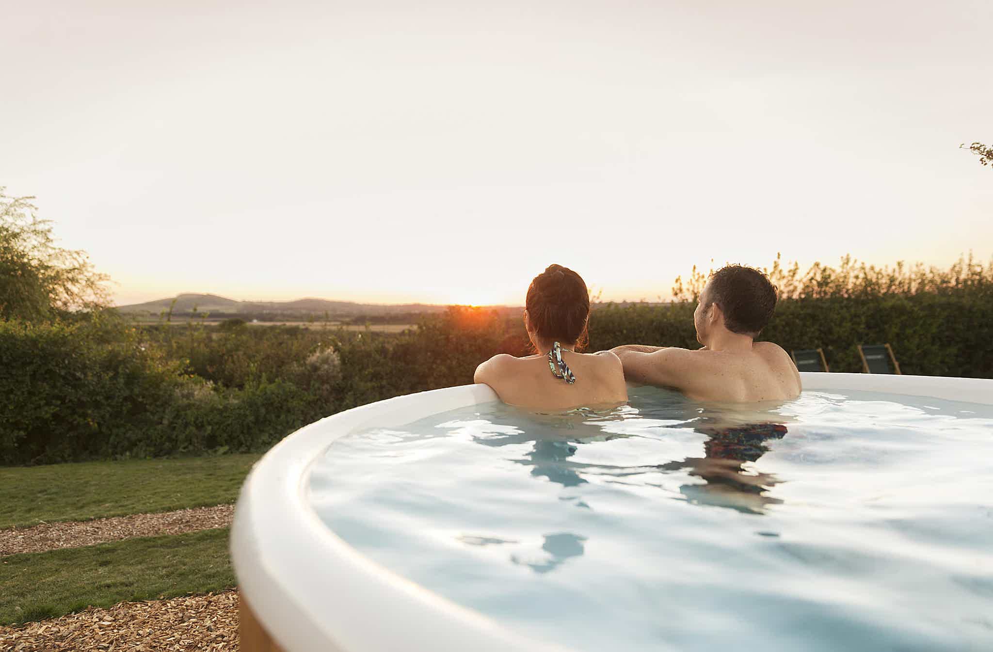 A couple relaxes in a Skargards hot tub amidst a garden, with hills and sunset in the background.