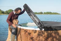 A man and child stand by a wooden hot tub on a dock. The tub has a black cover. The sky is clear.