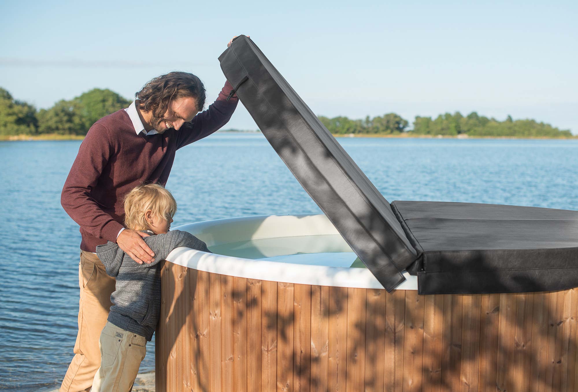 A man and child stand by a wooden hot tub on a dock. The tub has a black cover. The sky is clear.